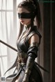 A woman in a dark outfit with a blindfold and a sword stands by a window.